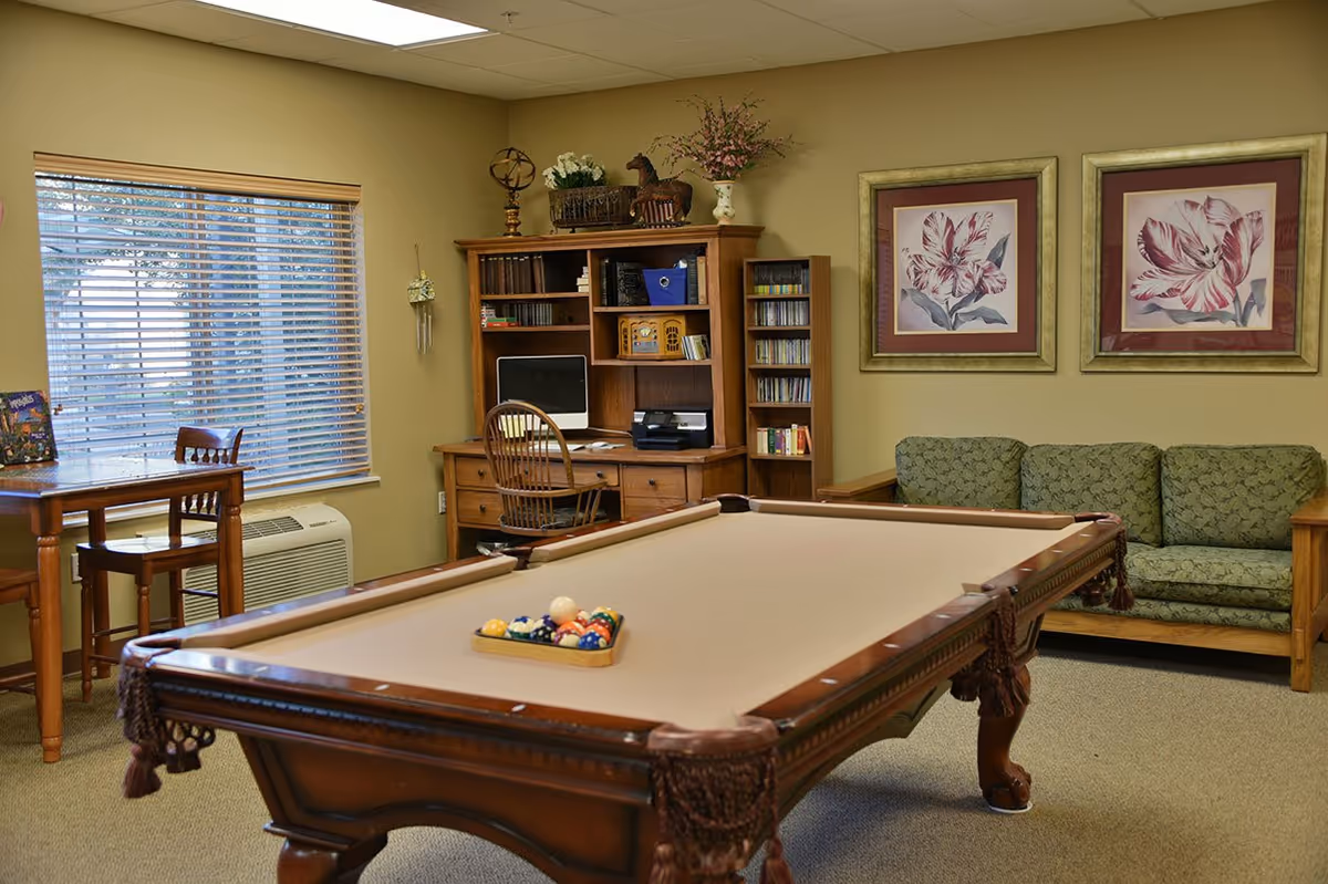 A cozy common room featuring a pool table with billiard balls arranged in a triangle, a wooden desk with a computer and chair, a small table with two chairs near a window with blinds, a green patterned couch, and two framed floral paintings on the wall.