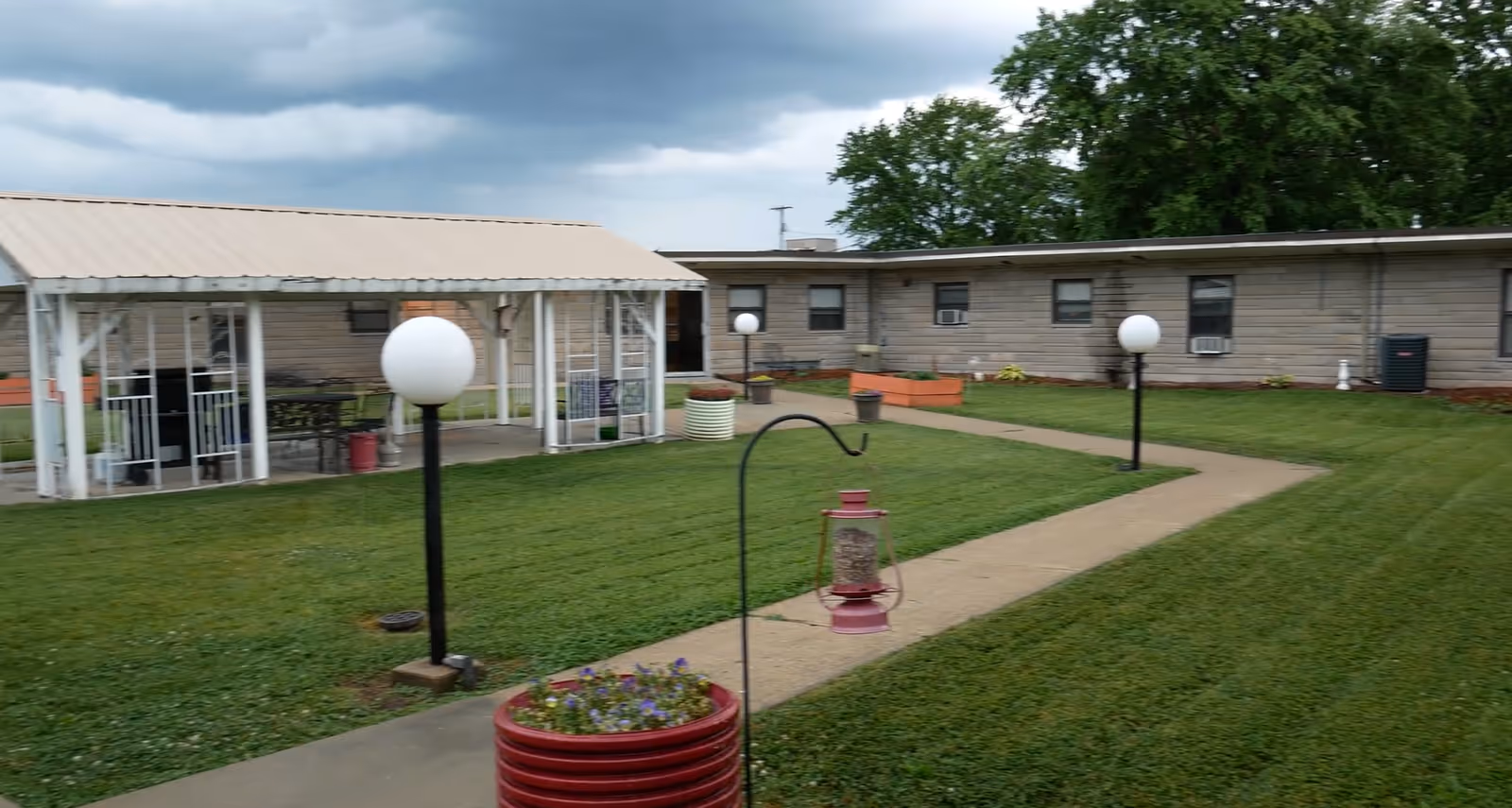 Outdoor courtyard area of a nursing and rehab facility with a covered seating area, green lawn, paved walkways, round white lamp posts, flower pots, and a single-story building in the background under a cloudy sky.