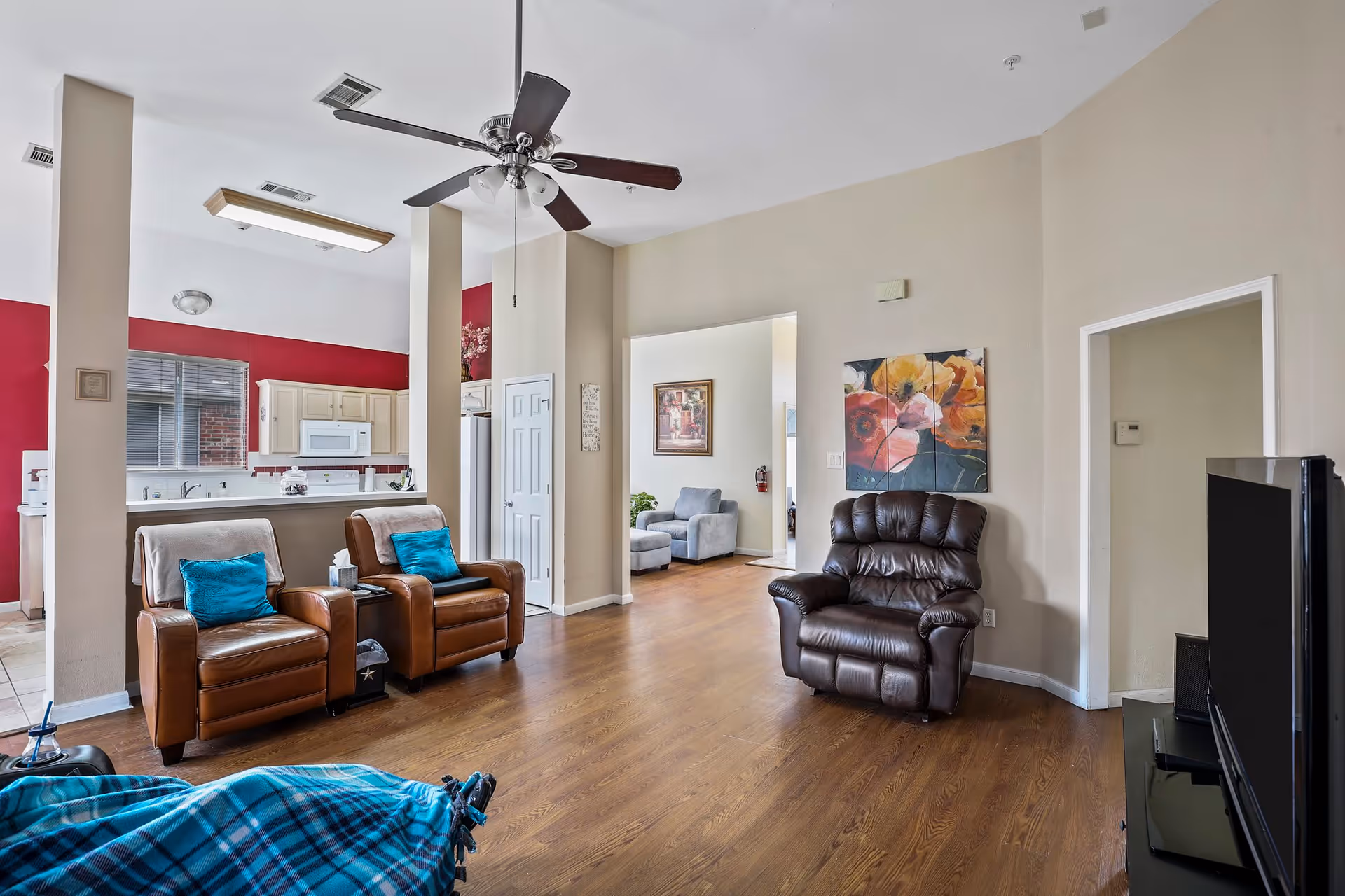 A spacious living room with wooden flooring featuring two brown leather armchairs with blue cushions, a dark brown recliner, and a flat-screen TV on a black stand. The room opens into a kitchen area with white cabinets and a red accent wall. A ceiling fan with lights hangs from the white ceiling. In the background, there is another seating area with a gray armchair and a framed picture on the wall.