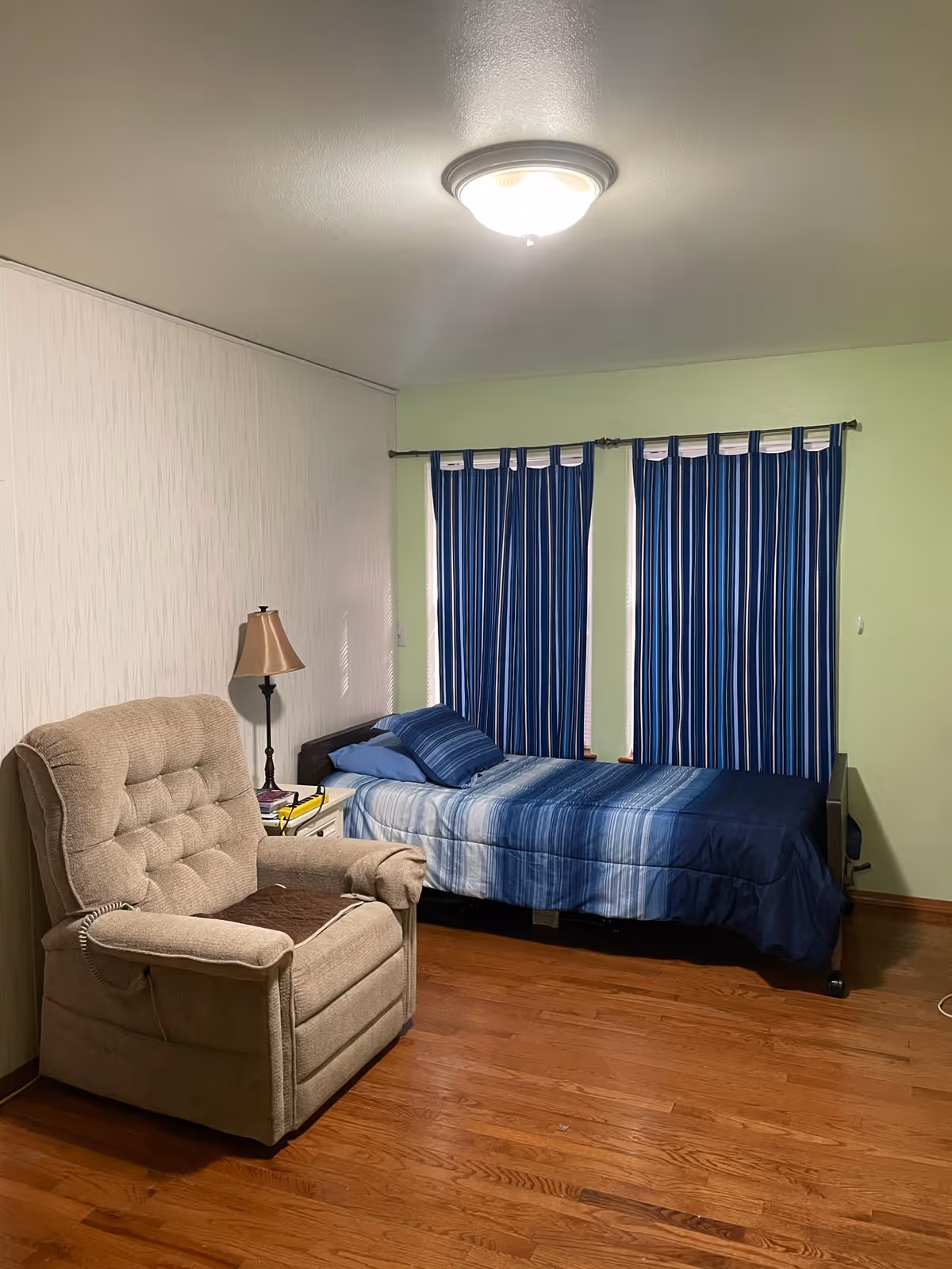 A bedroom with a single bed covered in blue striped bedding, two blue pillows, and a beige recliner chair. There is a small white nightstand with a lamp and some books next to the bed. The room has wooden flooring, light green walls, and two windows with blue and white striped curtains.