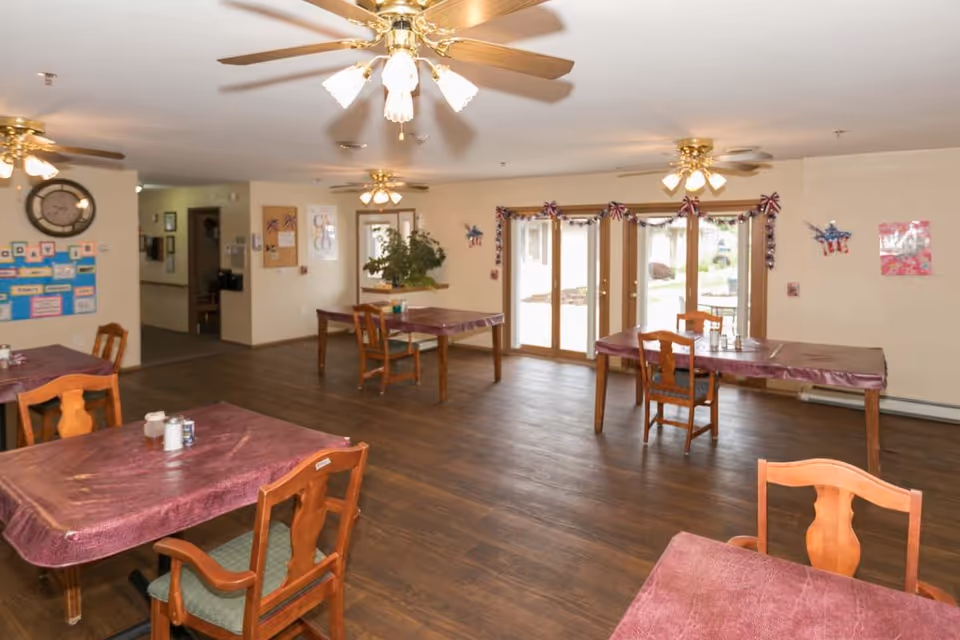 Spacious assisted-living dining room with several tables and chairs, ceiling fans, and large glass doors letting in daylight.