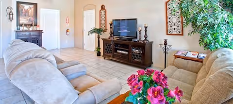 A bright living room with beige walls and tiled floor featuring two beige recliner chairs and a beige sofa. A wooden TV stand holds a flat-screen television and decorative items including candlesticks. There are framed artworks on the walls, a large green plant near the sofa, and a small table with a vibrant pink flower arrangement in the foreground.
