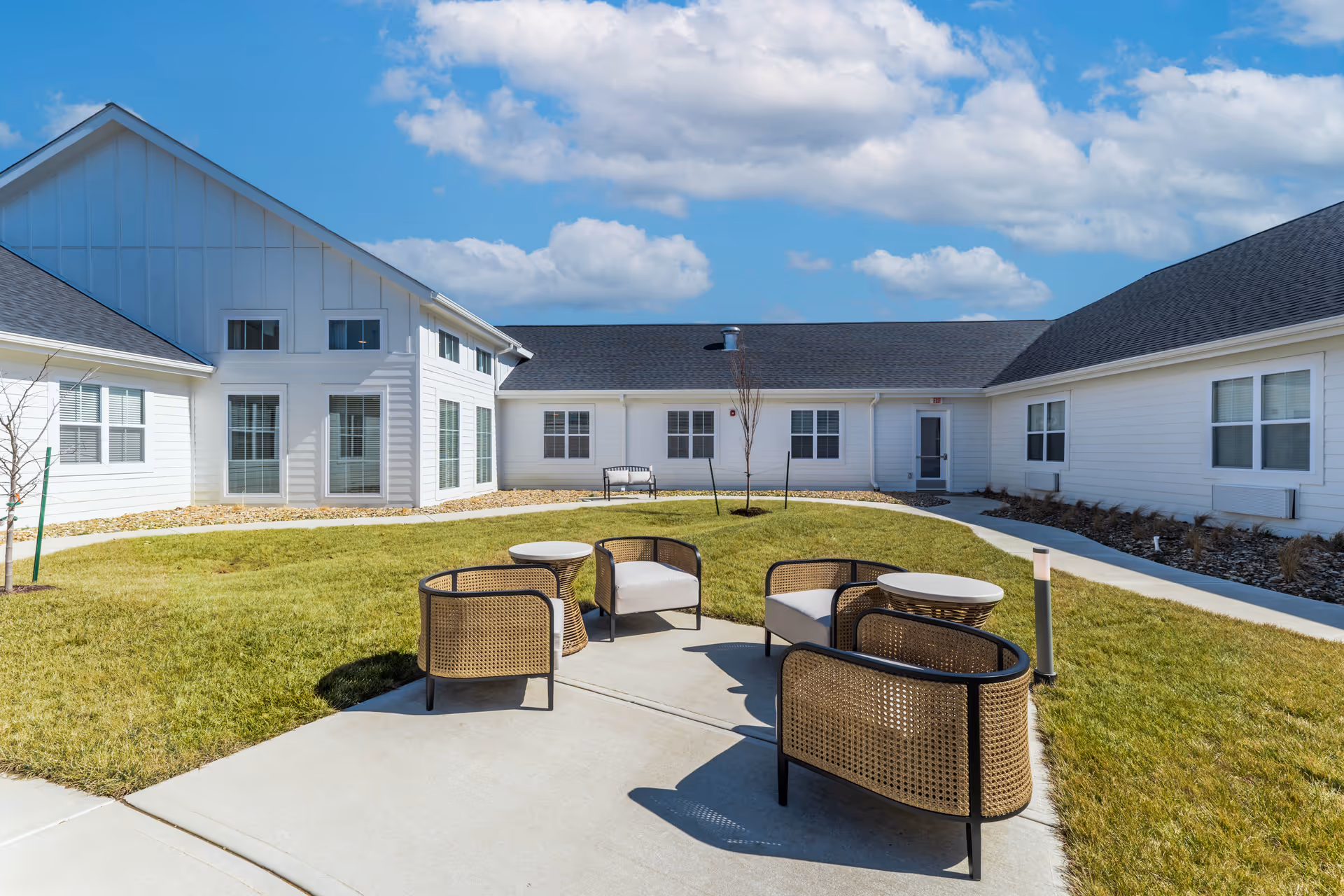 Outdoor courtyard area at Cedarhurst Senior Living of Topeka featuring a circular concrete patio with four wicker chairs and two small round tables, surrounded by green grass and white buildings under a blue sky with scattered clouds.