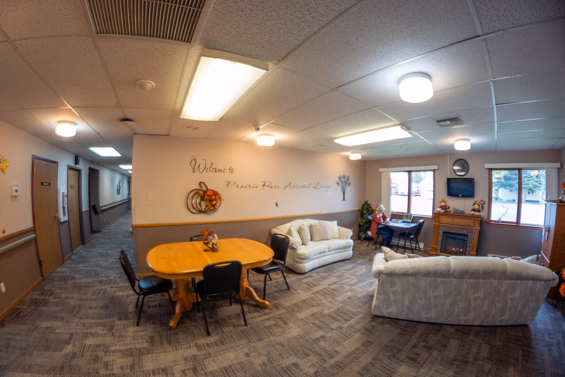 Interior view of a common area in Prairie Rose Assisted Living featuring two sofas, a wooden table with chairs, a fireplace, a wall-mounted TV, and a person sitting at a small table near the windows. The wall has a decorative sign that reads 'Welcome to Prairie Rose Assisted Living' with autumn-themed decorations.