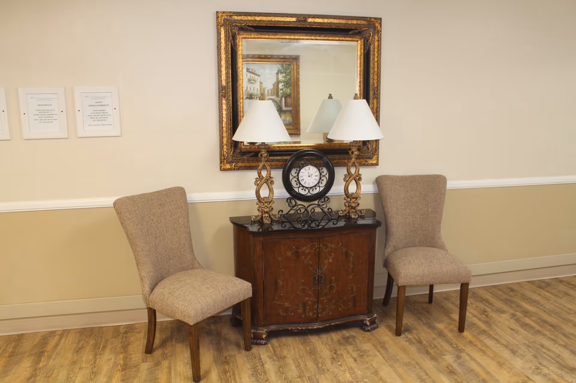 Two upholstered chairs flank a decorative wooden cabinet topped with a clock, lamps, and a framed mirror in a hallway-like interior.
