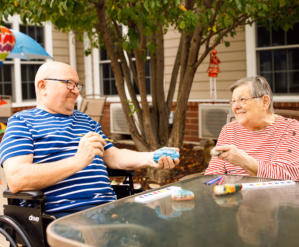 Two elderly individuals sitting outside at a glass table, painting rocks. One man in a blue and white striped shirt is holding a paintbrush and a blue rock, while a woman in a red and white striped shirt smiles at him holding a rock. They are seated near a tree with a building in the background.