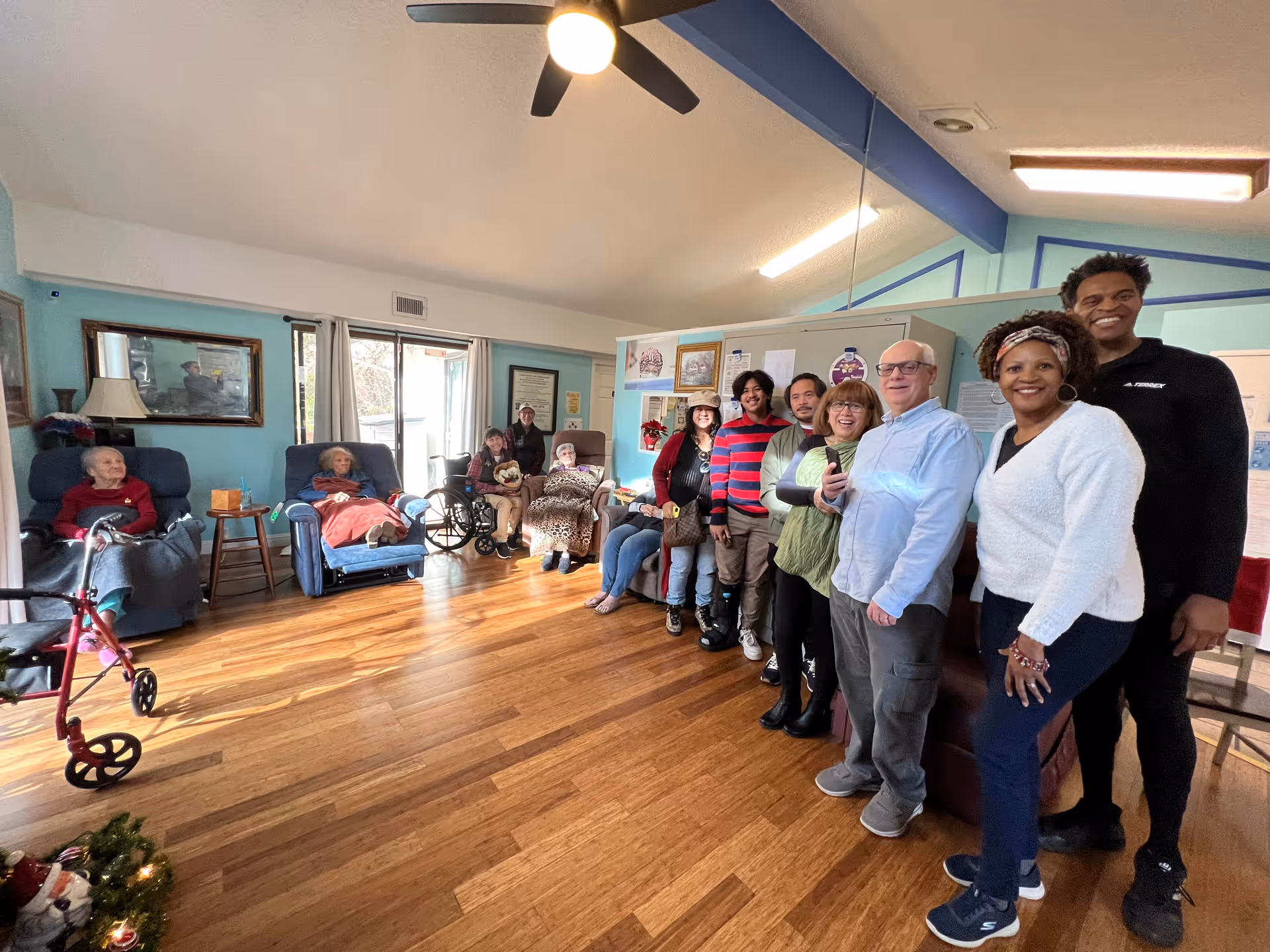 A group of elderly residents and staff members posing together in a spacious living room with wooden floors, light blue walls, and large windows letting in natural light. Some residents are seated in recliners and wheelchairs, while others stand smiling near a mirrored wall.