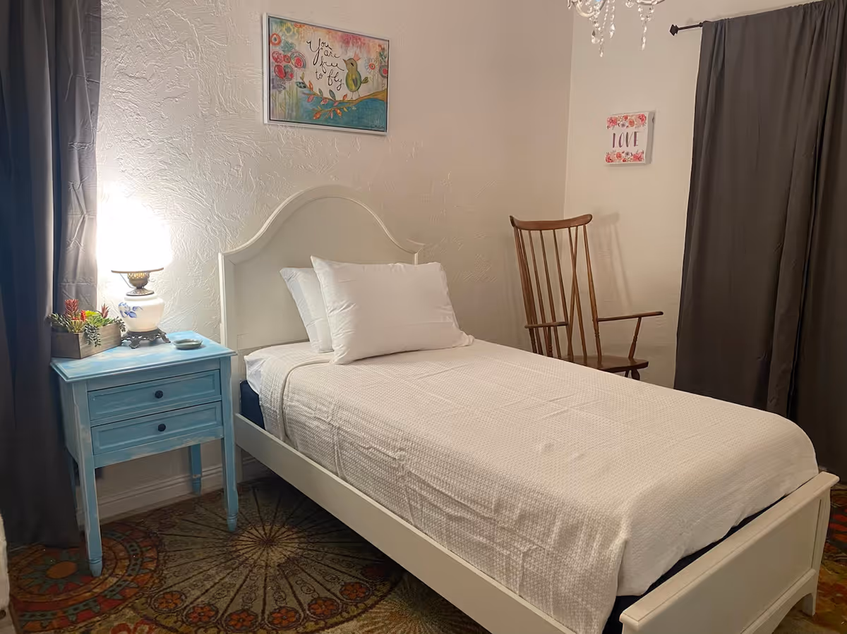 A cozy single bedroom with a white twin bed, blue nightstand and lamp, wooden rocking chair, and wall art on textured walls.