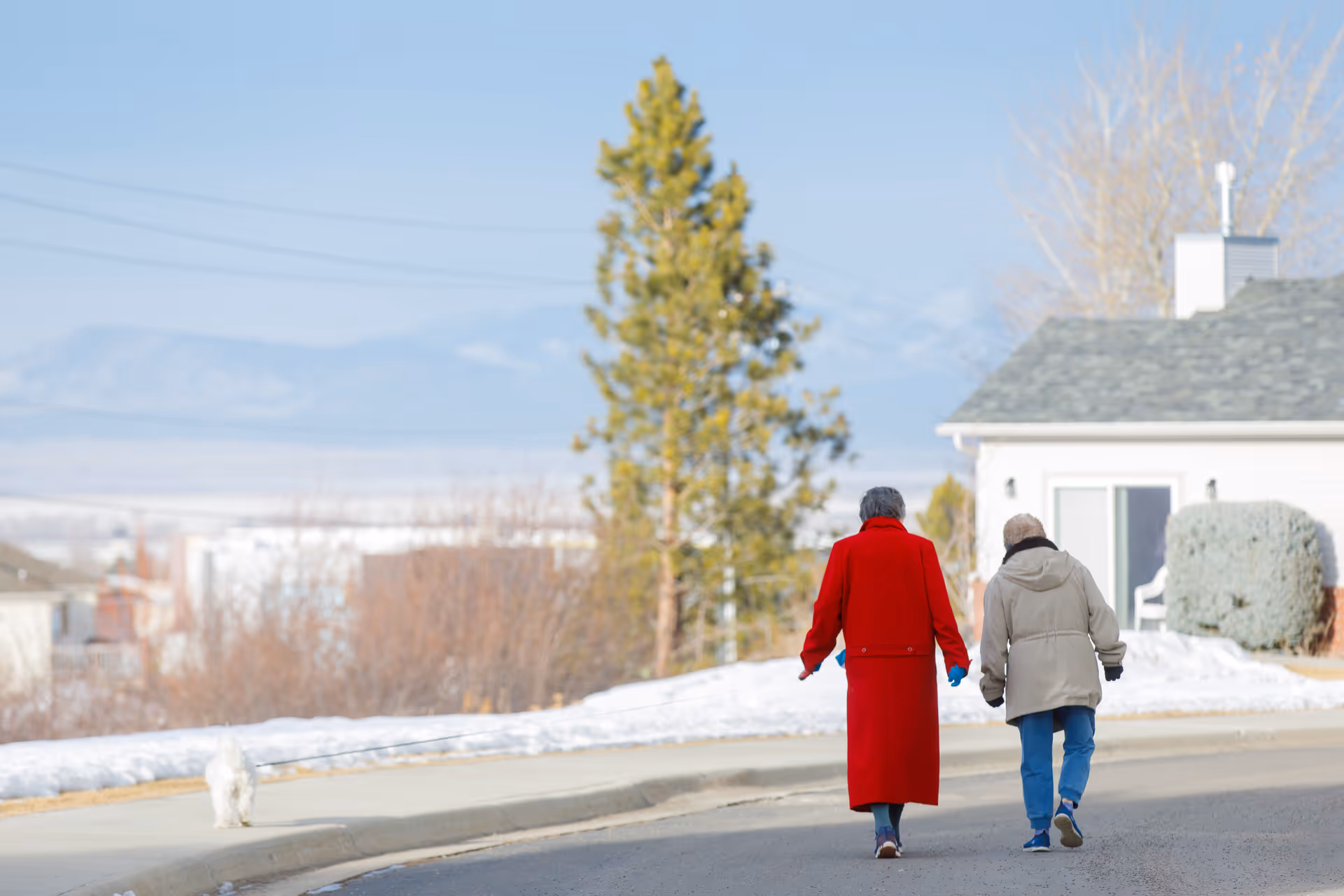 Two elderly people walking on a paved road in a residential area with snow on the ground. One person is wearing a long red coat and the other is wearing a beige jacket. A small white dog is walking on the sidewalk nearby. Trees, houses, and mountains are visible in the background under a clear blue sky.