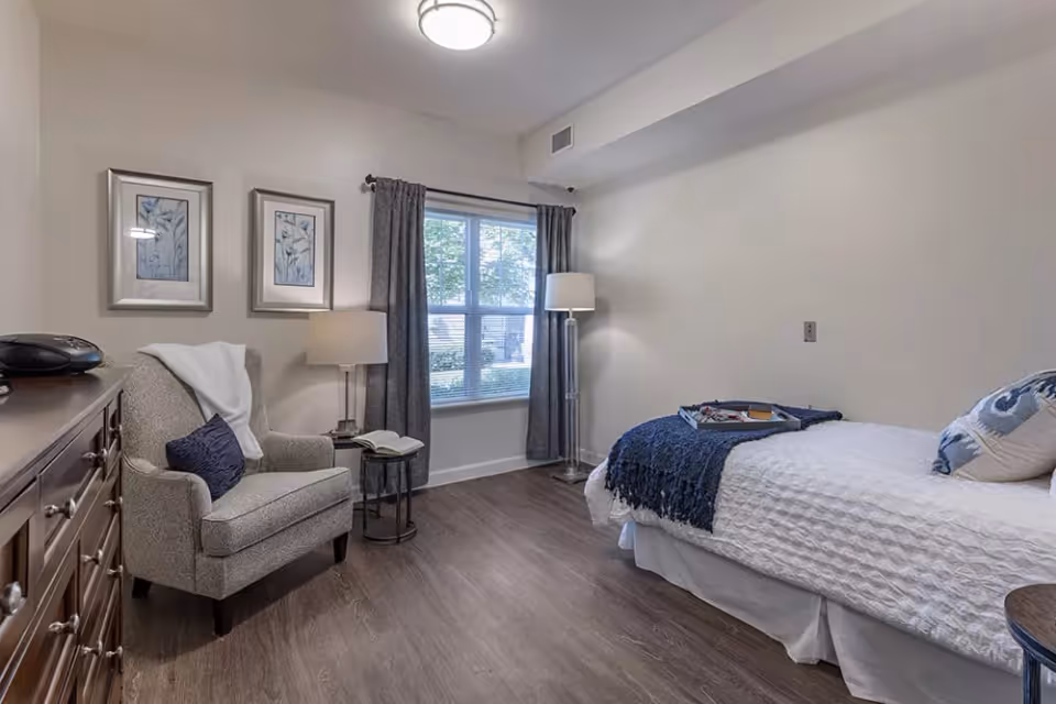 A cozy bedroom with a single bed covered in white bedding and a blue throw blanket. There is a cushioned armchair with a pillow and a white throw blanket next to a small round side table with an open book. A wooden dresser is on the left side of the room. The room has a window with gray curtains and two framed botanical prints on the wall. The floor is wood, and there is a ceiling light and a floor lamp near the bed.