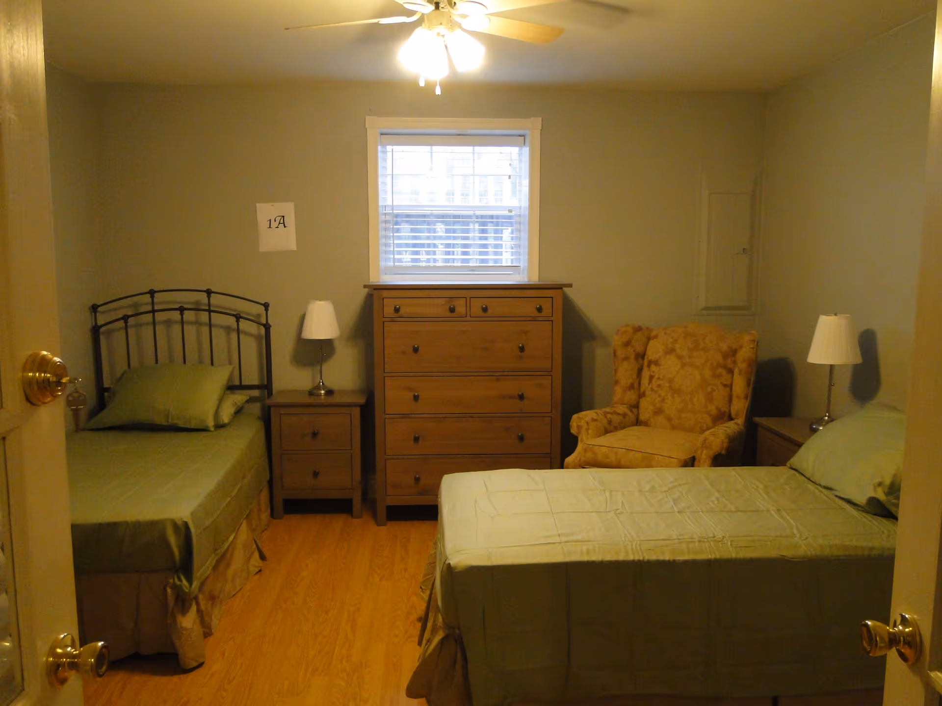 A small bedroom with two single beds covered in green bedding, a wooden dresser between the beds under a window with blinds, two nightstands each with a lamp, and a patterned armchair in the corner. The room has light-colored walls and a ceiling fan with lights.