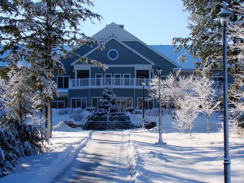 A large two-story building with a blue-gray exterior surrounded by snow-covered trees and ground. There is a pathway cleared of snow leading up to the building entrance, with steps and railings visible. The sky is clear and blue.
