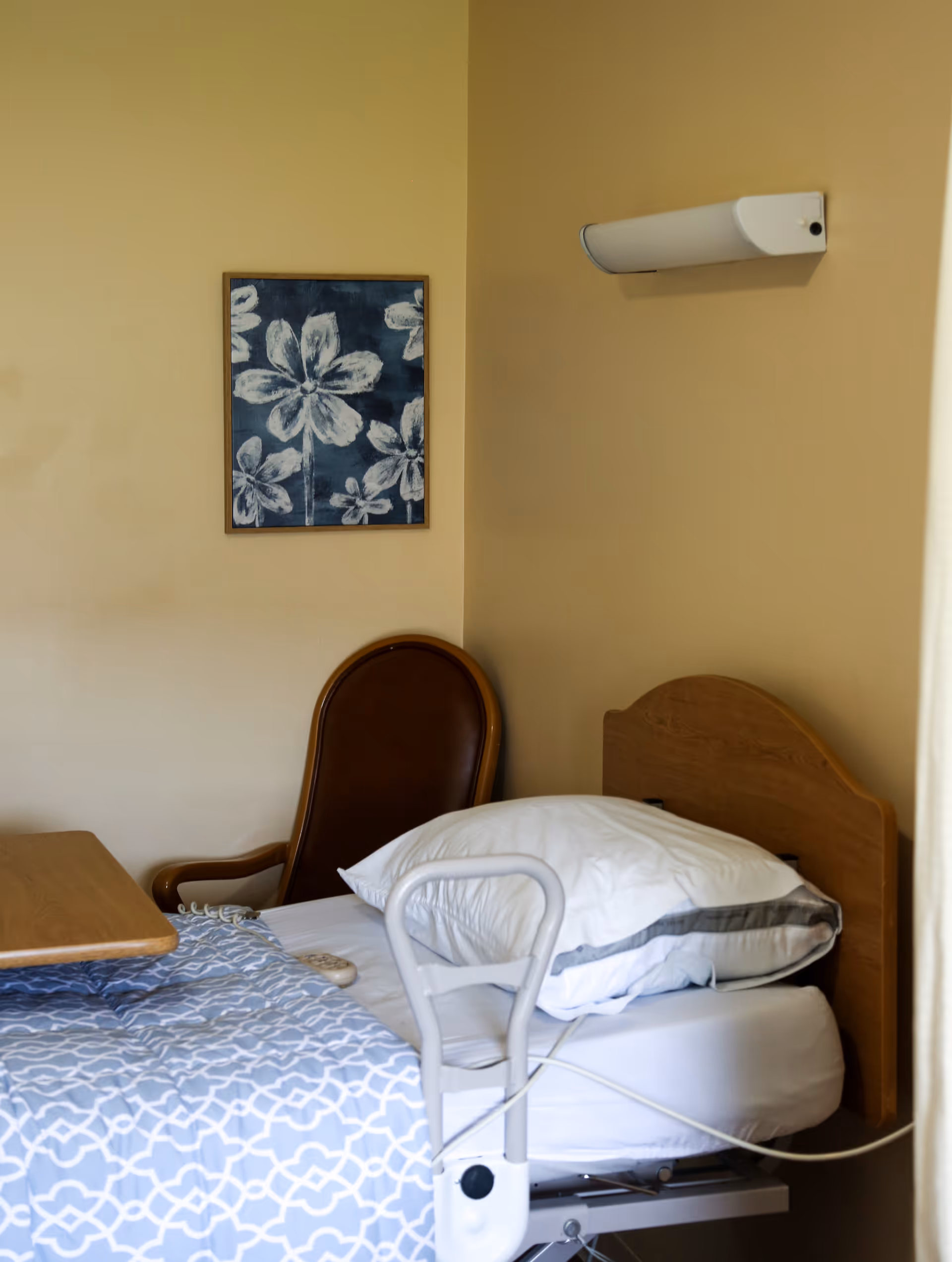 A hospital-style bed with white pillows and a blue patterned blanket in a room with beige walls. There is a wooden chair with a brown cushion next to the bed, a wooden headboard, a wall-mounted light fixture above the bed, and a framed floral artwork on the wall.