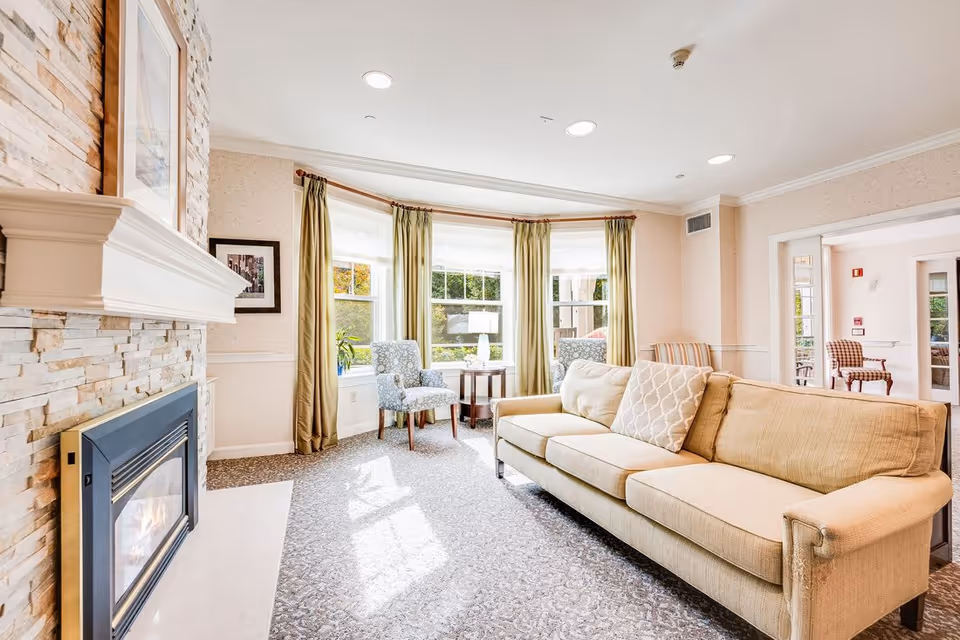A bright and cozy living room with a beige sofa, patterned armchairs, a stone fireplace with a white mantel, large windows with green curtains letting in natural light, and a small wooden table with a lamp between the chairs.