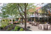 Outdoor patio area with several wooden tables and chairs arranged on a paved surface, surrounded by trees and landscaped garden beds. A white gazebo is visible on the left side, and a building with large glass windows is on the right.