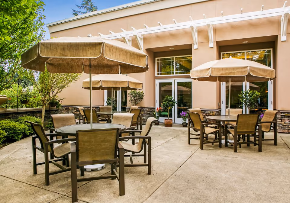 Outdoor patio with round tables, chairs and beige umbrellas in front of a building entrance.