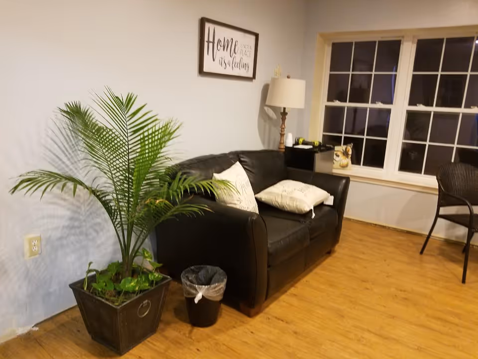 A cozy living room area with a black leather loveseat adorned with two white pillows. To the left of the loveseat is a large potted green plant and a small black trash bin. Behind the loveseat is a wooden floor lamp and a framed wall art that reads 'Home is not a place, it's a feeling.' There are two large windows on the right side with a small decorative item on the windowsill and a black chair nearby.