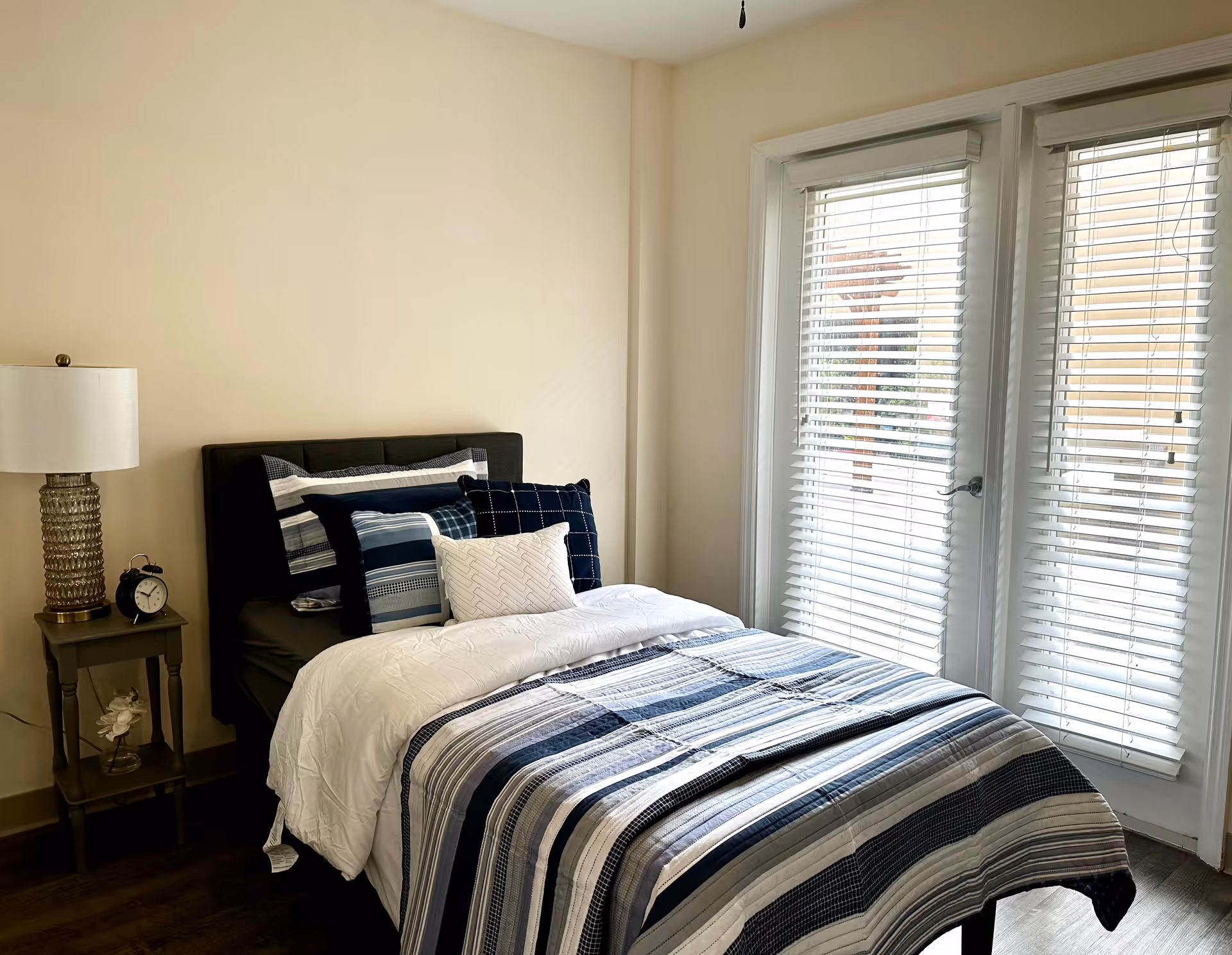 A bright bedroom with a single bed dressed in blue-and-white striped bedding, a nightstand with a lamp and alarm clock, and tall windows with blinds.