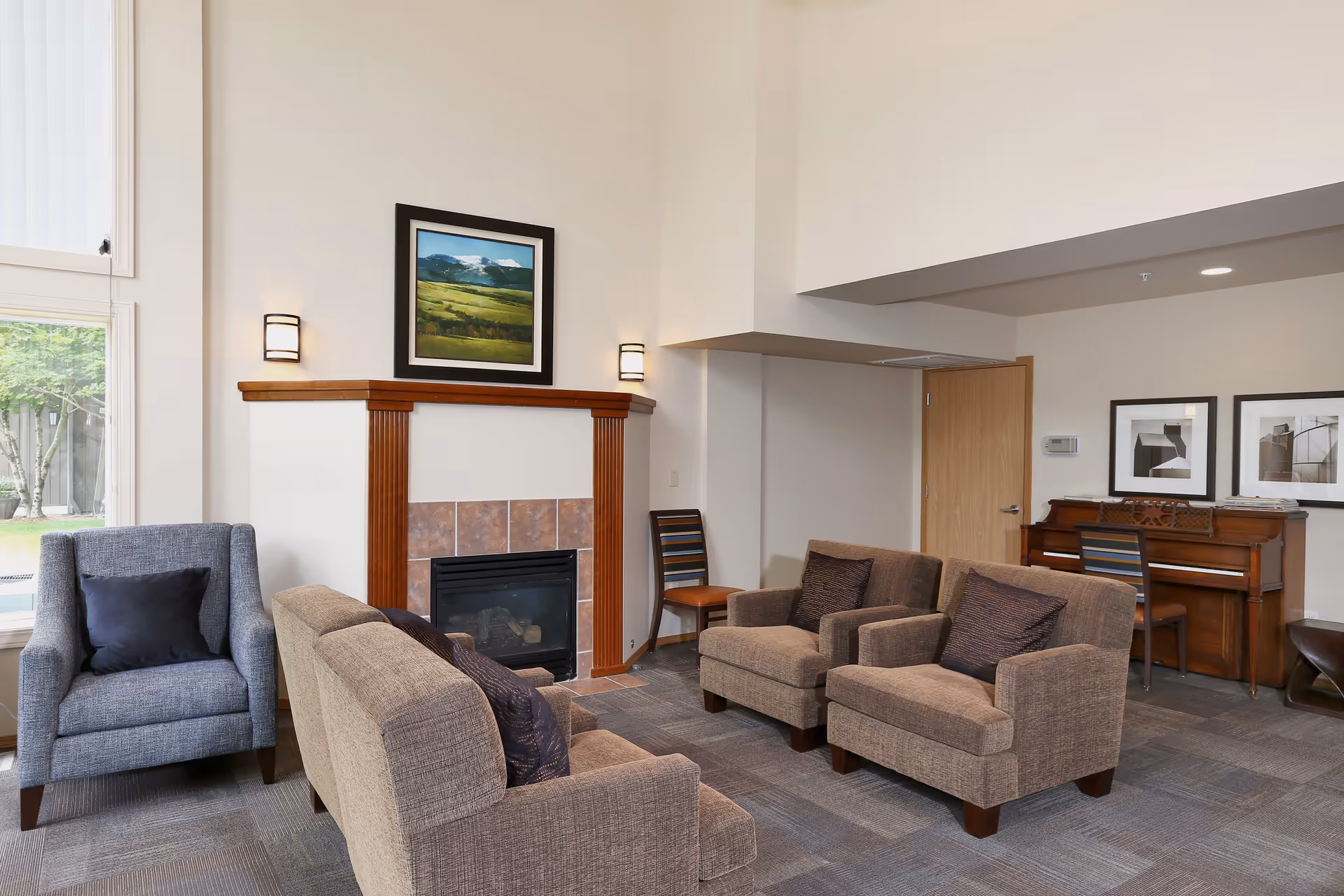 A cozy senior living room with a fireplace, two armchairs, a loveseat, a blue armchair with a black pillow, a wooden piano with a chair, and framed artwork on the walls. Large windows allow natural light to brighten the space.