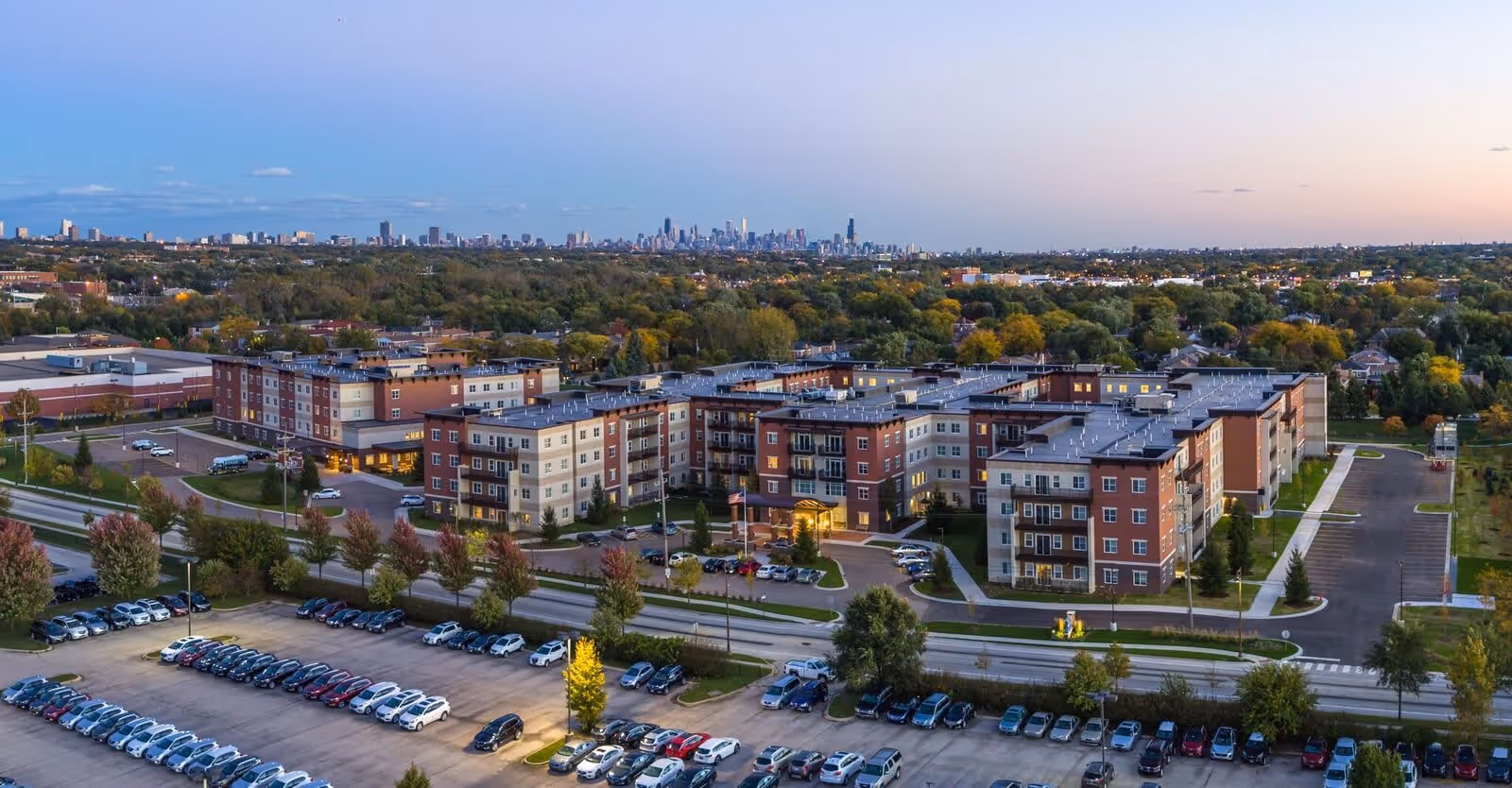 Aerial view of The Carrington at Lincolnwood, a large multi-story senior living facility surrounded by trees and parking lots, with a city skyline visible in the distance under a clear sky at dusk.