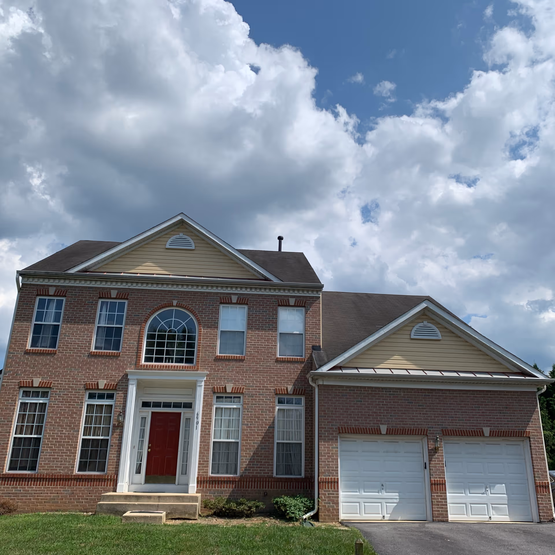 Two-story brick house with a red front door, attached two-car garage, and cloudy sky overhead.