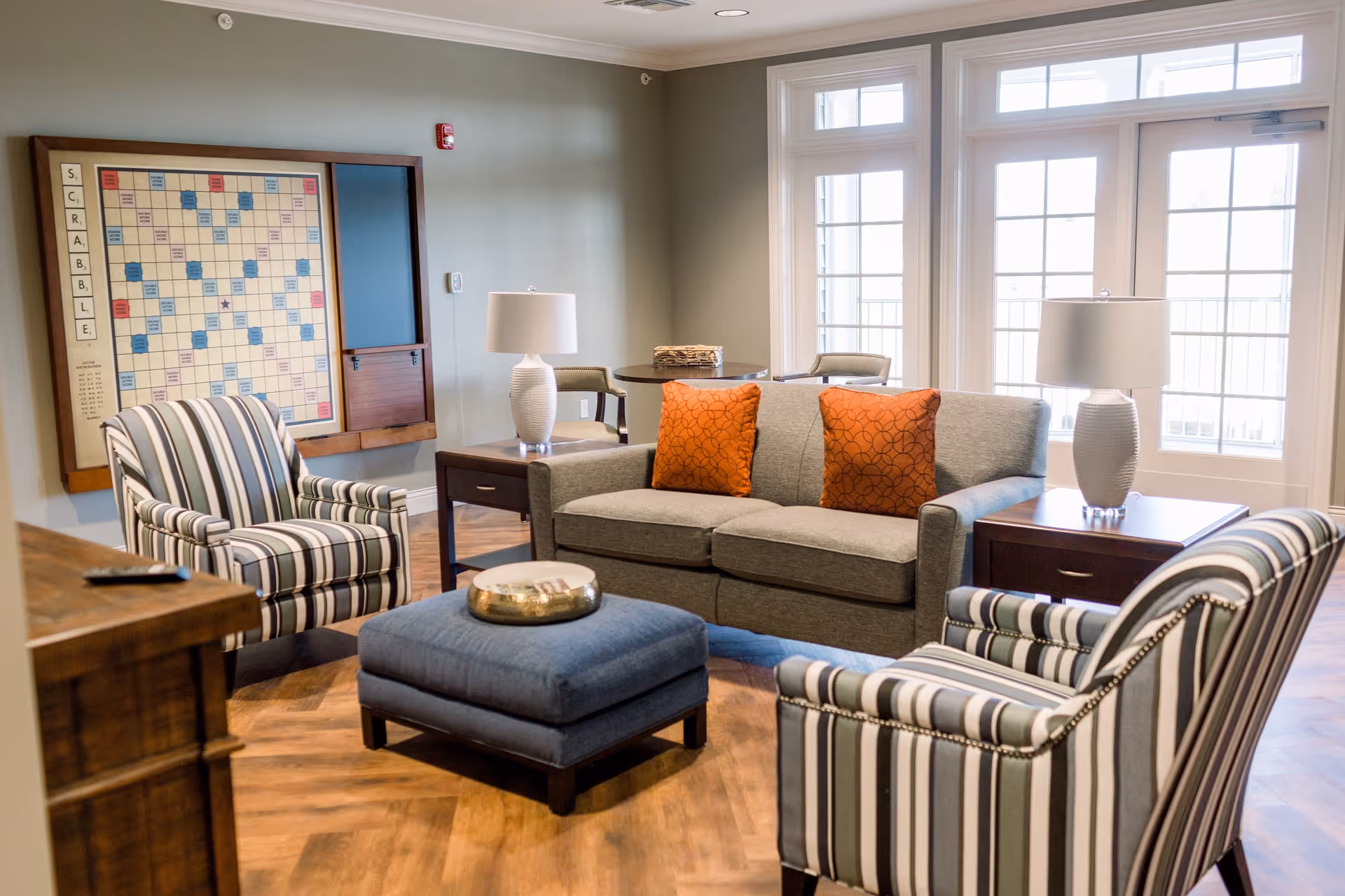 A cozy living room area in a senior living facility featuring a gray sofa with two orange patterned pillows, two striped armchairs, a blue ottoman with a decorative tray, two side tables with white lamps, and a large Scrabble board mounted on the wall. Large windows allow natural light to fill the room.