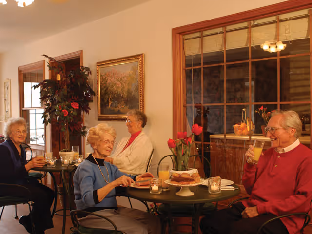 Four elderly residents seated at small round tables in a dining area enjoying drinks and pastries.