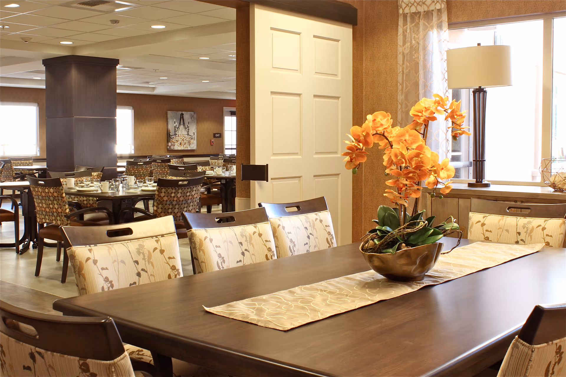 A well-lit dining area in a senior living facility featuring a large wooden table with floral-patterned chairs. An orange orchid centerpiece sits on a beige table runner. In the background, there are additional dining tables and chairs, a white sliding door, a floor lamp, and windows with sheer curtains allowing natural light to enter.