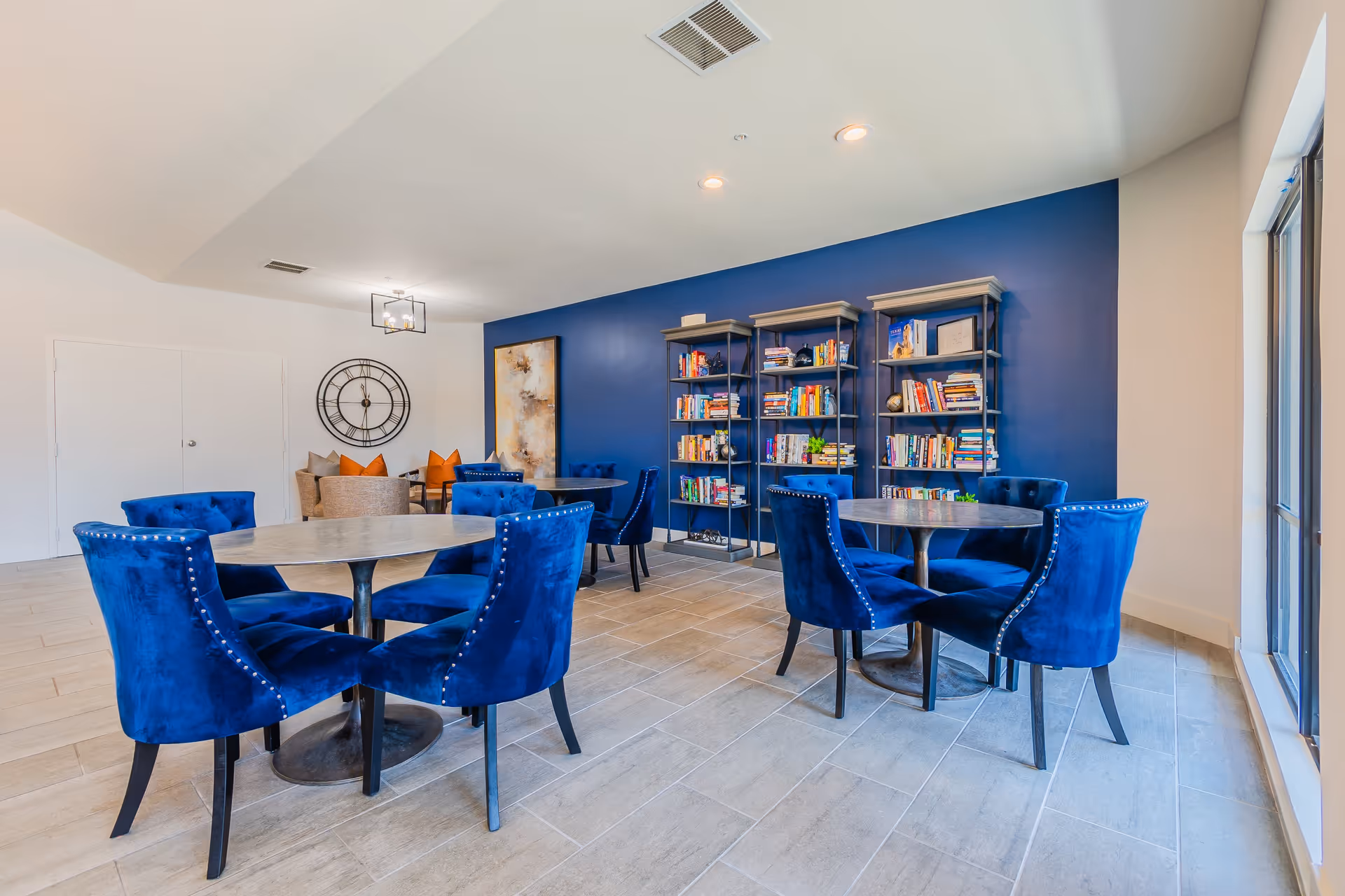 A bright and modern common area with two round tables surrounded by blue velvet chairs. In the background, there are three black metal bookshelves filled with books against a blue accent wall. A large decorative clock and a piece of abstract art are mounted on the adjacent white wall. The floor is tiled, and large windows on the right side let in natural light.