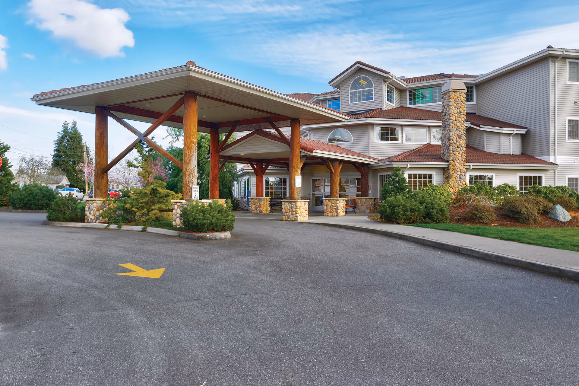 Exterior view of Creekside Continuing Care Community showing the main entrance with a covered drop-off area supported by wooden beams and stone bases, surrounded by landscaped greenery and a clear blue sky.