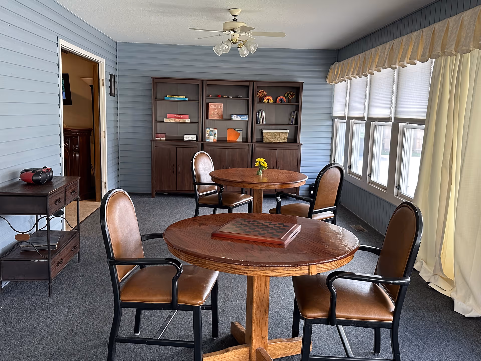 A cozy room with two round wooden tables, each surrounded by four brown leather chairs. One table has a chessboard on it and the other has a small vase with yellow flowers. There is a large window with white blinds and cream-colored curtains on the right side. A dark wooden bookshelf with books and decorative items is against the far wall. The walls are painted light blue with horizontal paneling, and there is a ceiling fan with lights above.