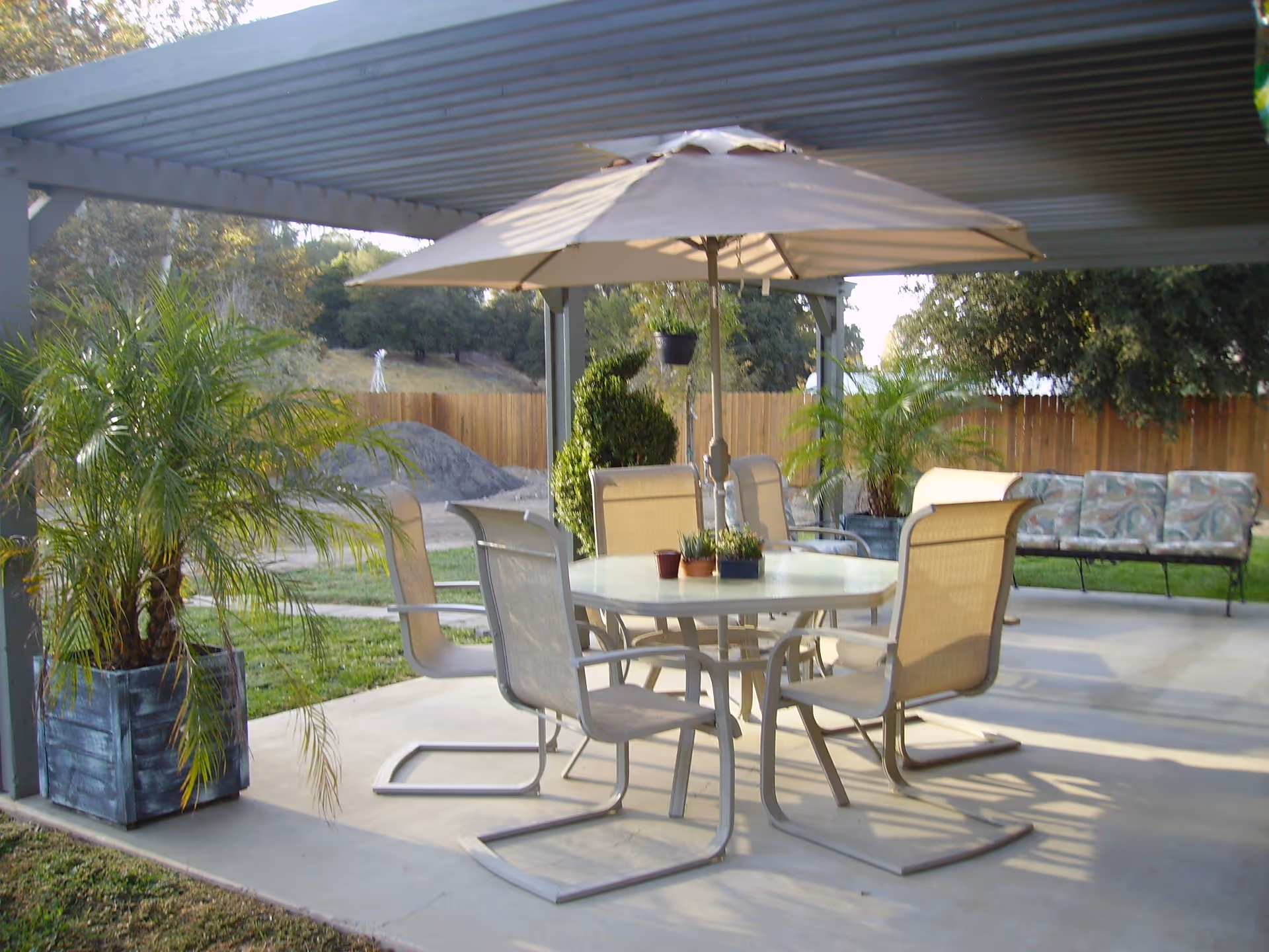 Covered backyard patio with a round table, umbrella and four chairs, potted plants and a bench under a pergola.