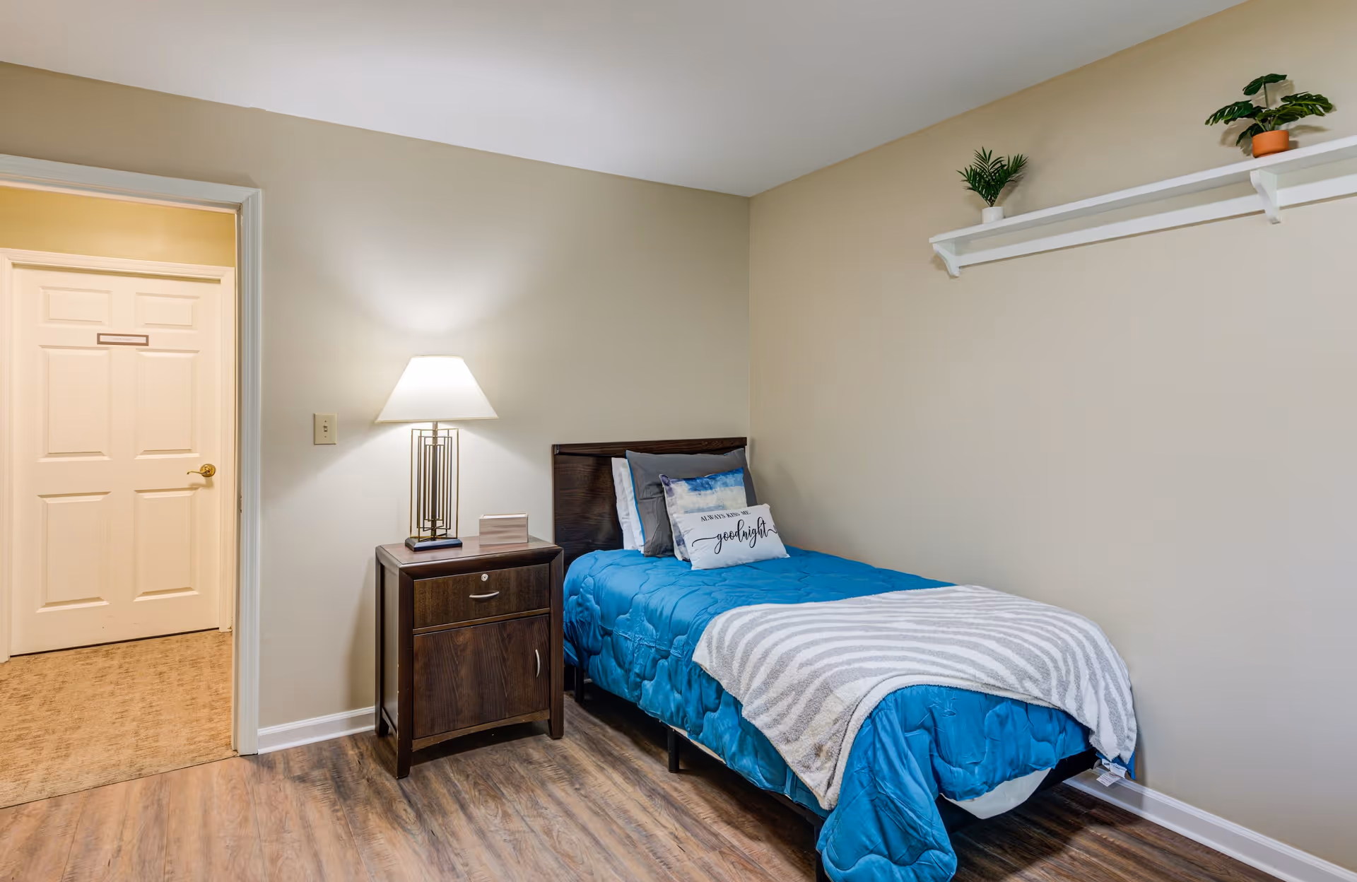 A small bedroom with a single bed covered in a blue comforter and a gray striped blanket. There are two pillows on the bed, one with a decorative design and the other with the word 'goodnight'. Next to the bed is a dark wooden nightstand with a lamp and a small decorative item. The walls are beige, and there is a white shelf with two small potted plants mounted on the wall above the bed. The floor is wood, and there is a doorway leading to another room with carpeted flooring.