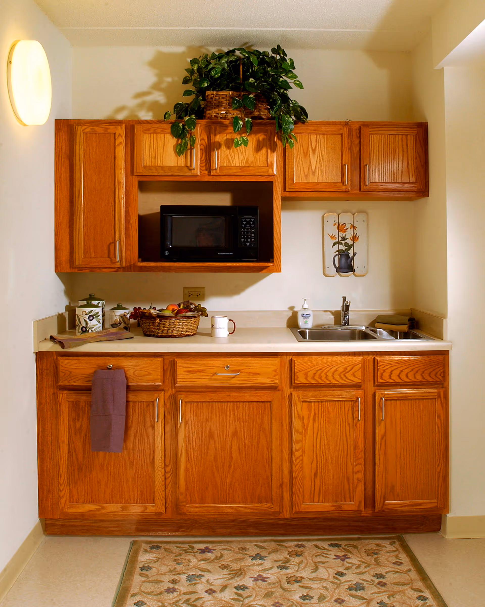 A small kitchen area with wooden cabinets, a countertop with a sink, a microwave oven, a basket of fruit, a mug, soap dispenser, and a decorative plant on top of the upper cabinets. There is a floral rug on the floor and a wall decoration with flowers above the sink.