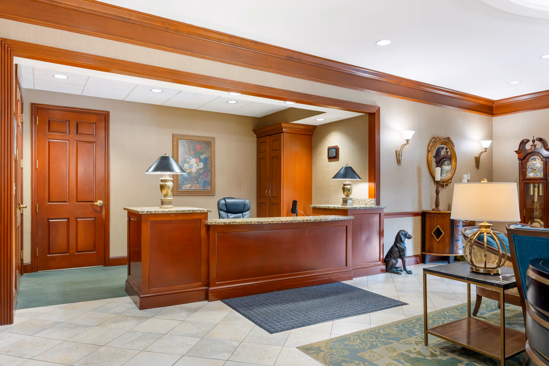 Reception area with a wooden front desk featuring two lamps with black shades, a black office chair behind the desk, a painting on the wall, and a wooden door to the left. The room has beige walls with wooden trim, a grandfather clock, a decorative mirror, wall sconces, a small table with a lamp, and a black dog statue sitting on the floor.