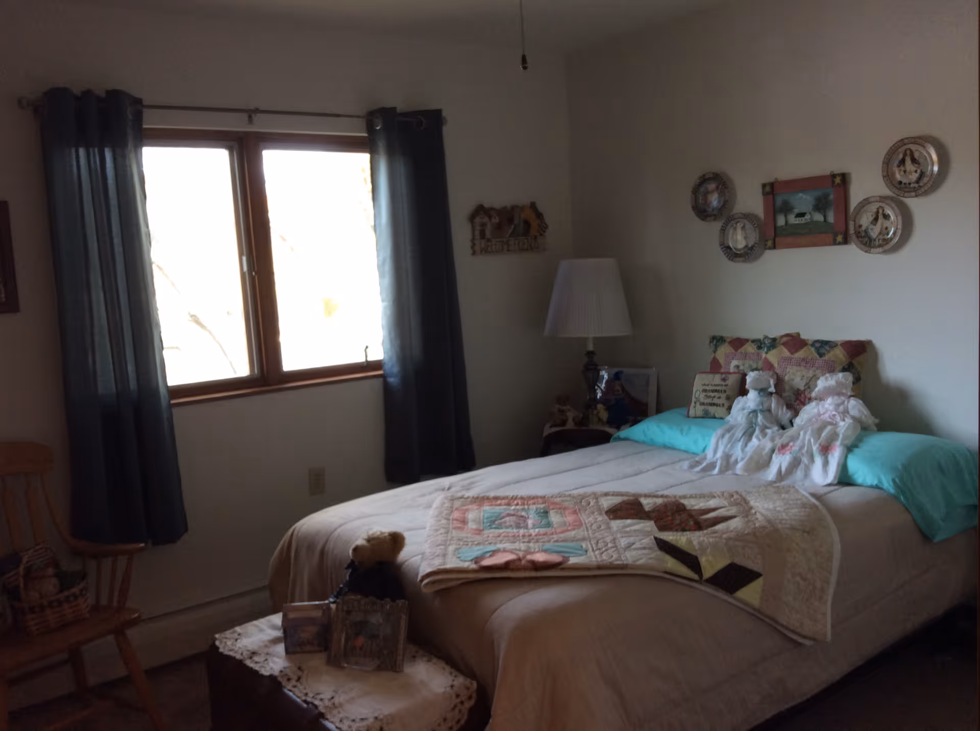 A cozy bedroom with a bed covered in a beige quilt and a colorful patchwork blanket. Two decorative dolls sit on the bed along with a pillow. The room has a window with dark curtains, a wooden chair with a basket, a small table with framed photos and a teddy bear, and a lamp on a side table. Decorative plates and a framed picture hang on the wall above the bed.