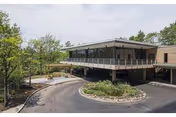 Exterior view of a modern senior living facility building surrounded by trees and landscaping, featuring a covered entrance with a circular driveway.