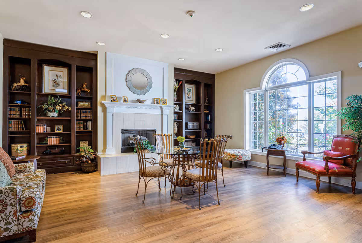 A bright and cozy living room with wooden flooring, a glass-top table with four metal chairs, a fireplace with a decorative mirror above it, built-in dark wood bookshelves filled with books and decorative items, a large arched window letting in natural light, a floral patterned sofa, a red upholstered armchair, and a small side table with a telephone.