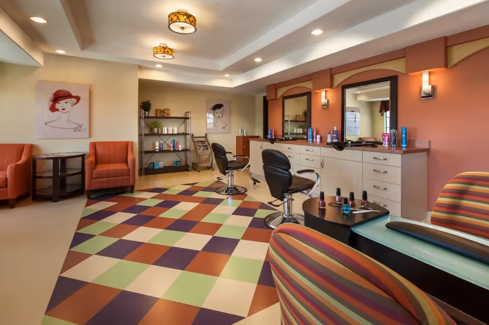 Interior view of a salon area in a senior living facility with two black salon chairs in front of mirrors and a counter with hair products. The room has colorful checkered flooring, striped armchairs, orange armchairs, and wall art featuring stylized portraits of women. The ceiling has recessed lighting and decorative light fixtures.
