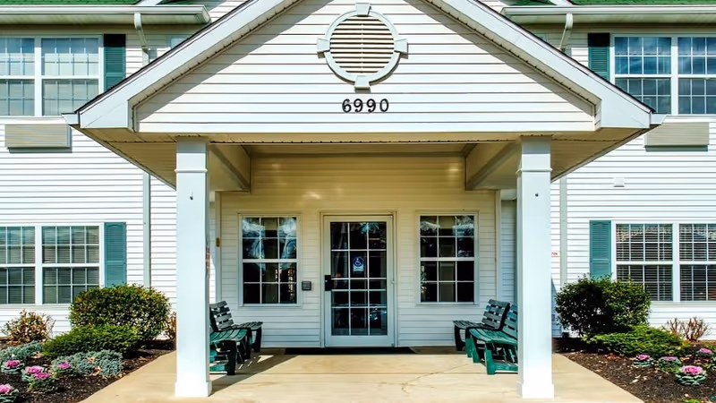 Covered front entrance of a two-story white building with benches, windows, landscaping, and the address number 6990 above the doorway.