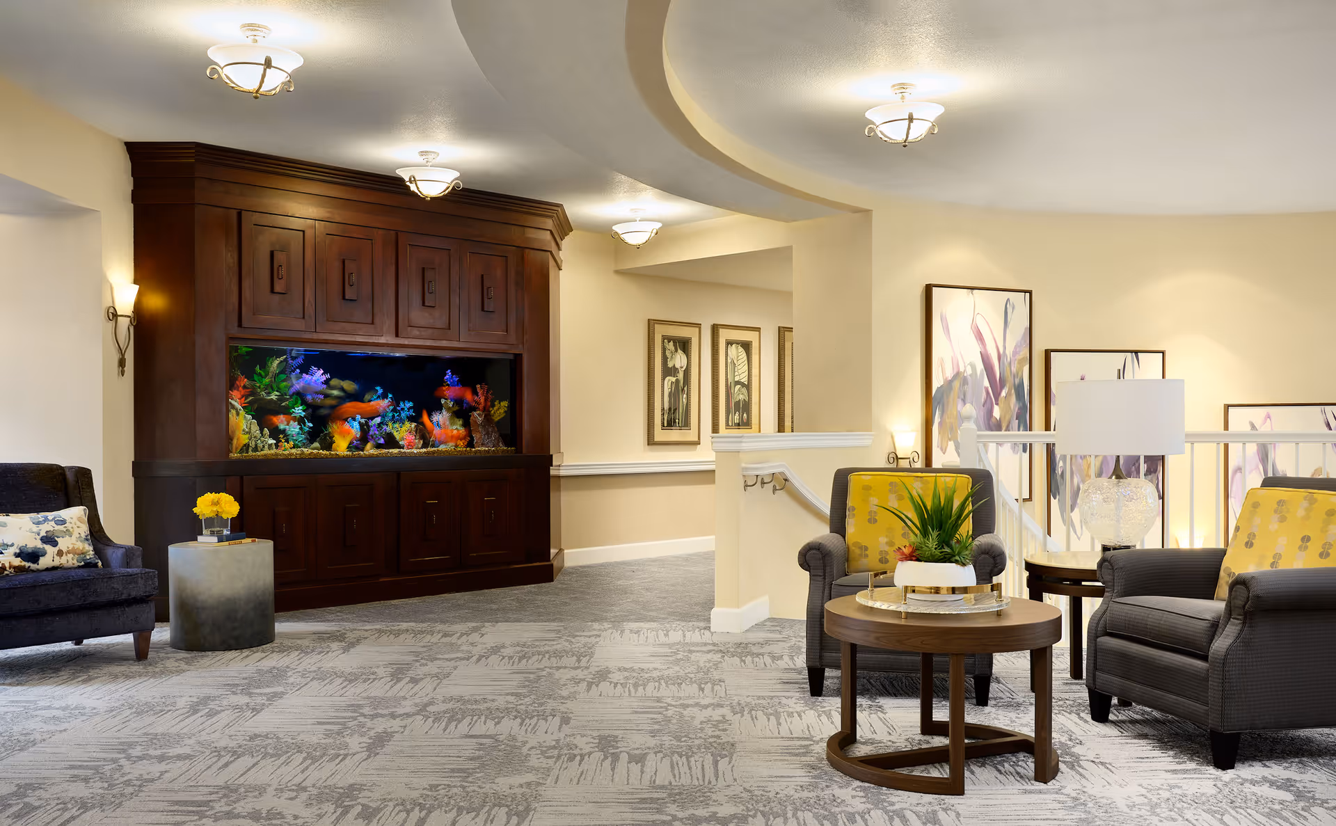 A well-lit interior common area featuring a large built-in aquarium with colorful fish and coral, surrounded by dark wood cabinetry. The space includes comfortable seating with two gray armchairs adorned with yellow patterned cushions, a round wooden coffee table with a plant centerpiece, and a side table with a lamp. The walls are decorated with framed artwork and the carpet has a textured pattern.