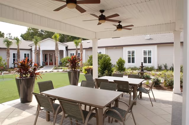Covered outdoor patio area with ceiling fans, several tables and chairs, large potted plants, and a view of a garden with palm trees and a building in the background.