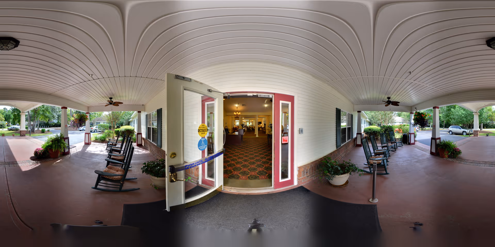 Covered entrance area of a senior living facility with several rocking chairs lined up on both sides, potted plants, ceiling fans, and an open glass door leading inside to a carpeted lobby area with warm lighting.