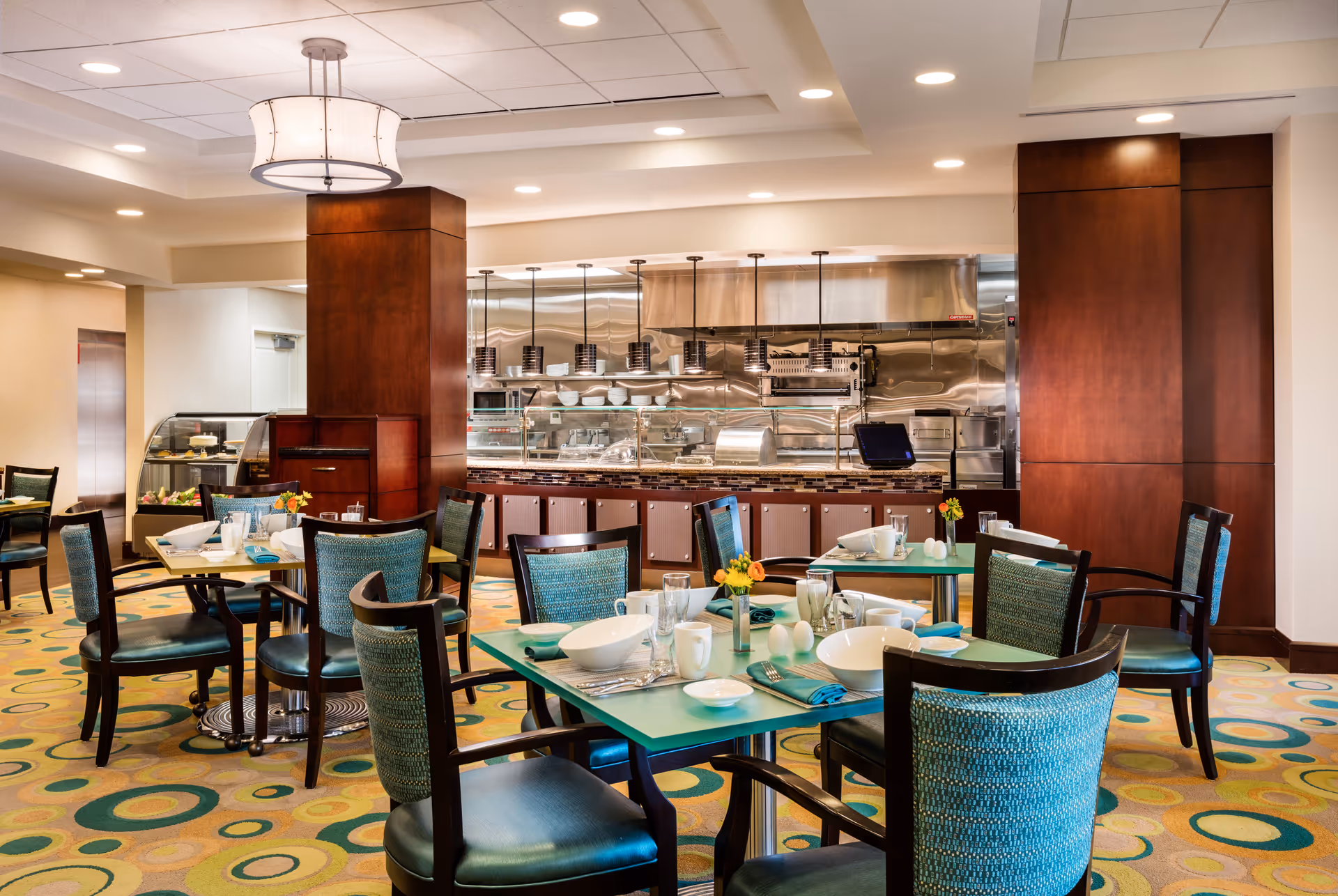 A bright and modern dining room with teal and dark wood chairs around tables set with white bowls, plates, cups, and silverware. The room features a patterned carpet with circular designs and a large light fixture hanging from the ceiling. In the background, there is a serving counter with stainless steel kitchen equipment and a glass display case with desserts.