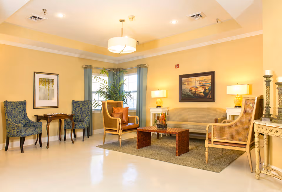 A cozy living room area with beige walls and a light-colored floor. The room features a beige sofa, two wicker armchairs with cushions, a wooden coffee table on a gray rug, and two side tables with yellow lamps. There are two blue patterned chairs with a small wooden table between them near a window with blue curtains. The walls are decorated with framed artwork, and there are decorative candles on a small ornate table to the right.