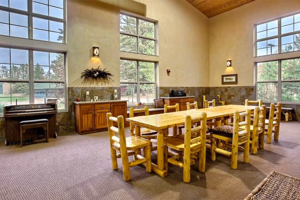 Spacious senior living common room with a long rustic wooden dining table and chairs under tall windows and a high ceiling.