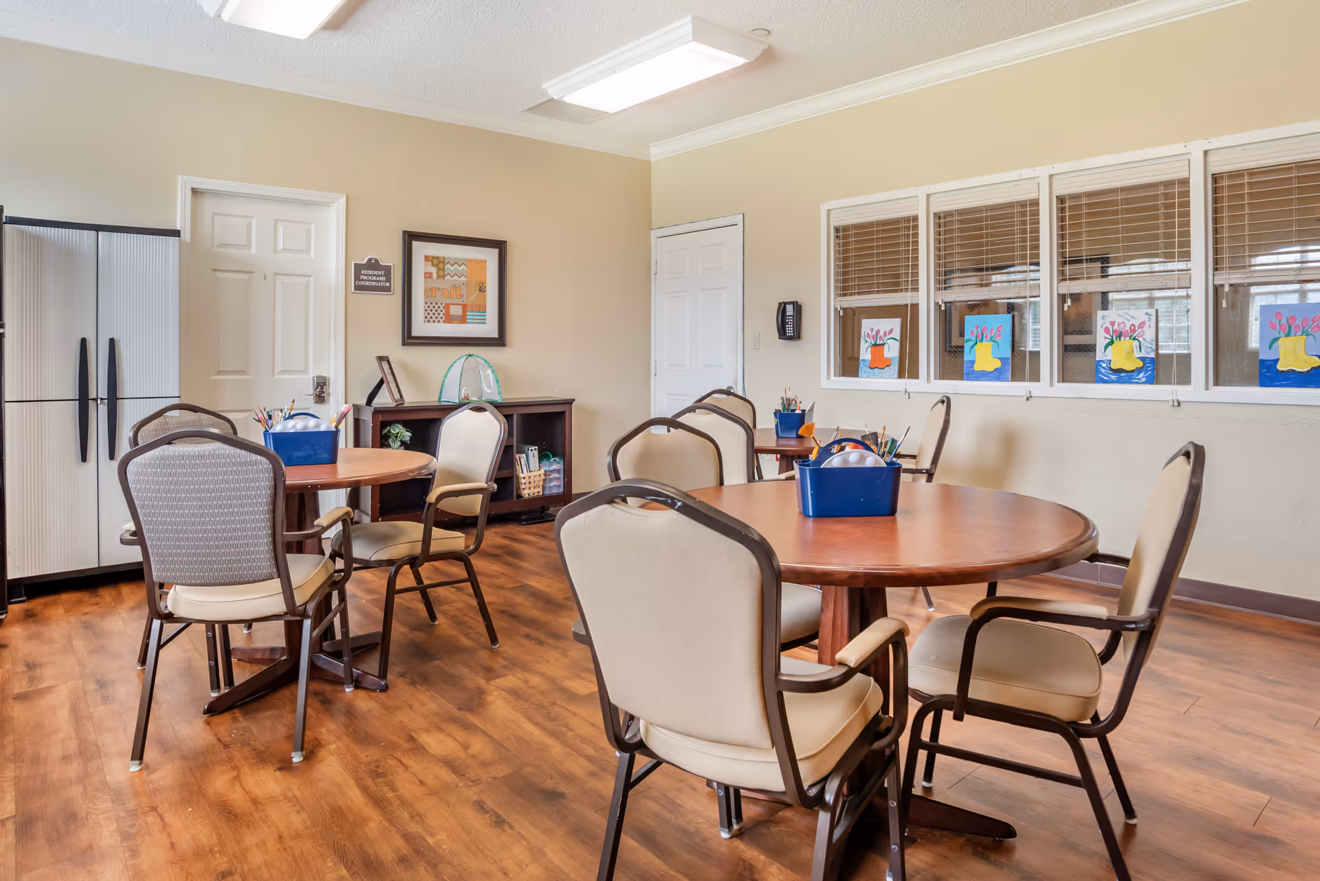 A well-lit activity room with round wooden tables and beige cushioned chairs arranged around them. Each table has a blue container filled with art supplies like paintbrushes and colored pencils. The room has wooden flooring, beige walls, and a set of windows with blinds displaying colorful flower paintings. There is a door labeled 'Resident Programs Coordinator' and a cabinet against the wall.