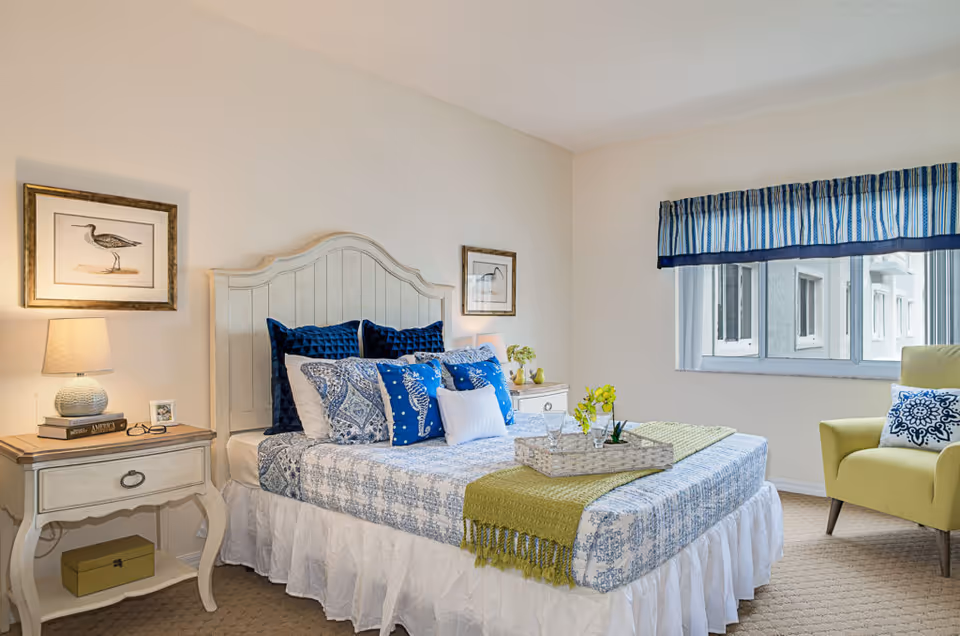 A neatly made bed with a white wooden headboard, decorated with multiple blue and white pillows and a green throw blanket. On the left side, there is a nightstand with a lamp, books, and a small framed photo. On the right side, there is another nightstand with a small plant. A window with blue and white striped valance is on the right wall, next to a green armchair with a decorative pillow. The room has beige carpet and light-colored walls.