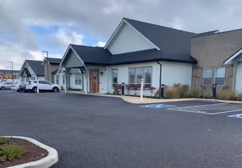 Exterior view of a single-story building with multiple peaked roofs and a parking lot in front, including handicap parking spaces. Several cars are parked along the side of the building under a cloudy sky.
