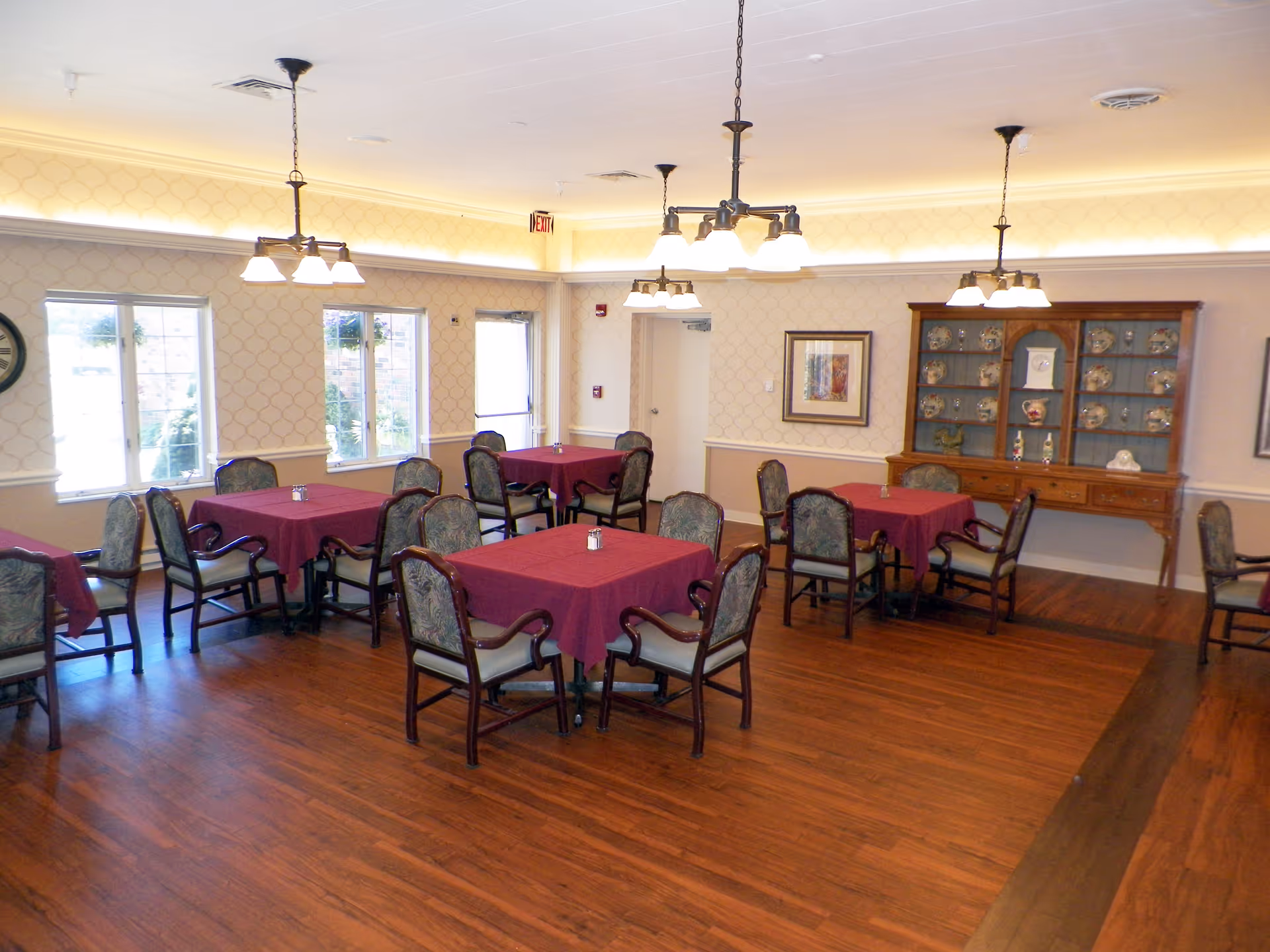 A dining room with several tables covered in burgundy tablecloths, each surrounded by four upholstered chairs. The room has wooden flooring, patterned wallpaper, and multiple hanging light fixtures. There is a wooden cabinet displaying decorative plates and items against one wall, and windows letting in natural light.